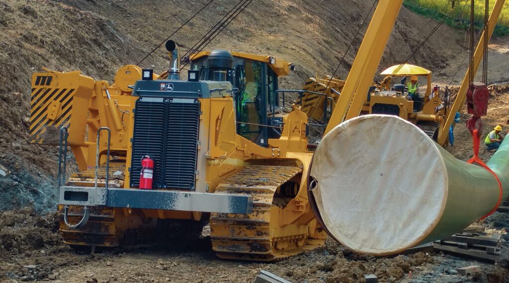 Heavy yellow pipeline sideboom crawler tractors lowering a large insulated pipeline section into a trench at an active construction site, with workers in high-visibility safety gear visible in the background.