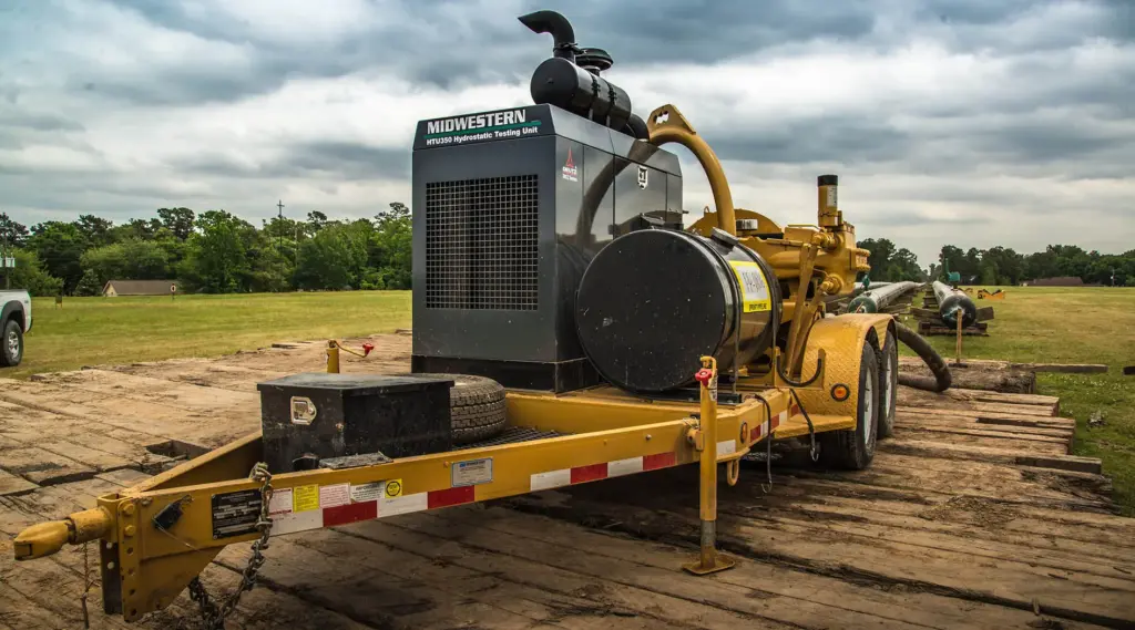 Midwestern HTU350 hydrostatic testing unit mounted on a yellow trailer at an outdoor pipeline testing site, with pipeline sections and additional equipment visible in the background.